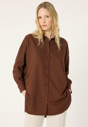 Femme blonde portant une chemise marron ample à manches longues et boutonnée, les mains dans les poches, debout devant un fond clair uni.