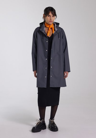 Grijze waterdichte regenjas met een capuchon, drukknoopsluitingen en koordjes. G gedragen over een zwarte jurk en gecombineerd met dikke zwarte schoenen. Oranje sjaal om de nek.