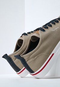 Zapatillas de lona beige con cordones negros, suelas de goma y acentos rojos. Presentan un parche de cuero marrón en la lengüeta. Costura alternante visible.