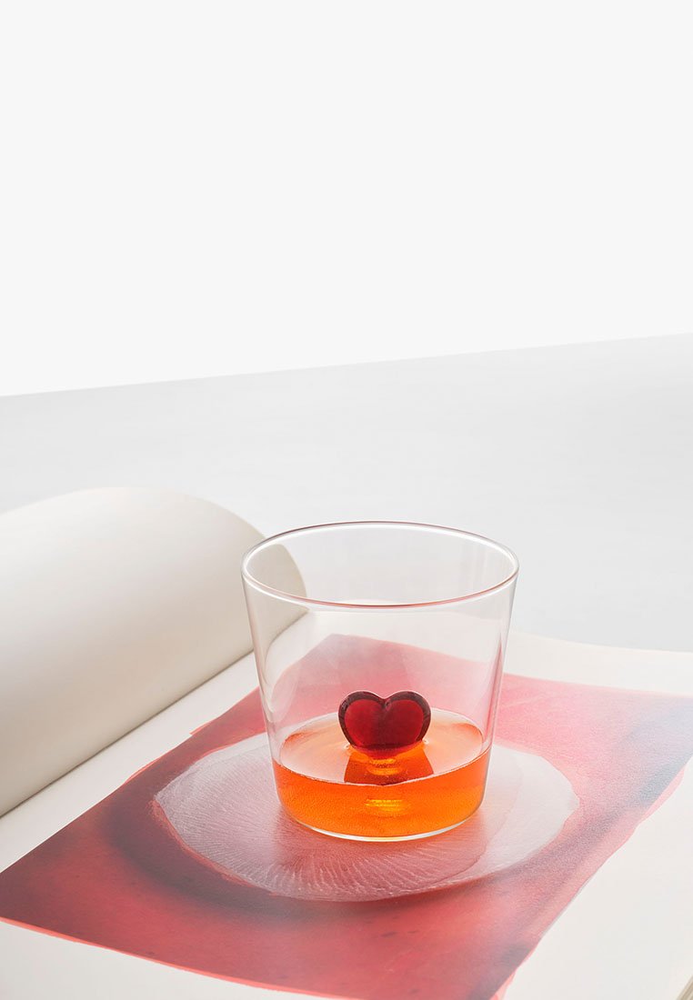 Durchsichtiges Glas mit einem roten herzförmigen Objekt darin, das auf einer bunten Kunstbuchseite mit abstrakten Mustern ruht.