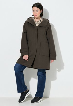 Woman wearing a dark brown hooded coat over plaid shirt, blue jeans, and black sneakers standing against a plain light background.
