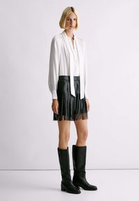 Witte lange mouw blouse met een kraag, gecombineerd met een zwarte leren fringes minirok en zwarte laarzen met blokhak tot de knie.