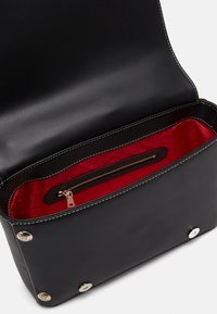 Svart läderhandväska med en slät yta, röd inredning och en ficka med dragkedja. Guldiga detaljer på v äskans botten.