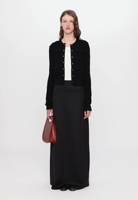 Cardigan nero con bottoni e due tasche a patta, indossato su una maglietta bianca. Gonna nera lunga, abbinata a una borsa marrone.