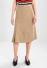 Lichtbeige A-lijn rok van een gladde stof met een zijknopdetail, met minimale stiksels en een halflange zoom. Gecombineerd met zwarte hakken.