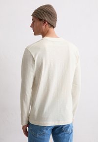 Junger Mann mit einer braunen Strickmütze, einem weißen Langarmshirt und blauen Jeans, der vor einer schlichten weißen Wand steht und nach links schaut.