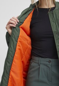 Prešívaná zelená bomber bunda s oranžovým interiérom, ktorá má zips a okrúhly výstrih, vrstvená cez čiernu košeľu a zelené nohavice.