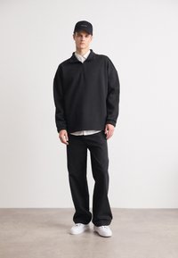 Pullover in pile nero e oversize con zip a un quarto, abbinato a pantaloni neri a vestibilità rilassata e sneakers bianche, completato da un cappellino nero.