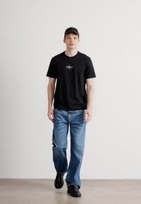 T-shirt preta de manga curta com um logotipo central, jeans de ganga azul clara e sapatos pretos. O modelo está a usar um boné preto correspondente.