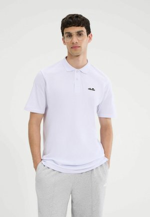 Młody mężczyzna w przezroczystych okularach, białej poloszortce z logo i jasnoszarych dresach, stojący z rękami w kieszeniach.