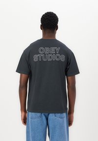 Melns kokvilnas T-krekls ar īsām piedurknēm, ar baltu uzrakstu "OBEY STUDIOS", kas drukāts uz muguras. Klasisks apaļš kakla izgriezums.