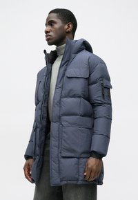 Φουσκωτό μπουφάν σε σκούρο ναυτικό χρώμα, με κουκούλα, πολλές τσέπες και υφή. Με φερμουάρ και κλείσιμο με κουμπιά, σε μοντέρνα γραμμή.