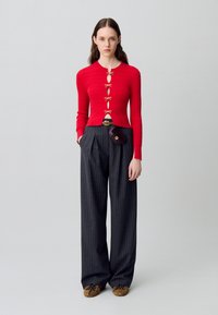 Maglioncino rosso a coste con dettagli in oro a forma di fiocco e tagli, abbinato a pantaloni a vita alta con righe sottili e scarpe stringate marroni.