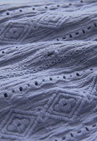 Světlomodrá texturovaná látka s vystouplými diamantovými vzory a malými otevřenými stehy, vytvářející jemný, detailní povrch.