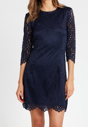 Mujer que lleva un vestido de encaje azul marino con borde en forma de ondas y mangas tres cuartos, sobre un fondo blanco liso.