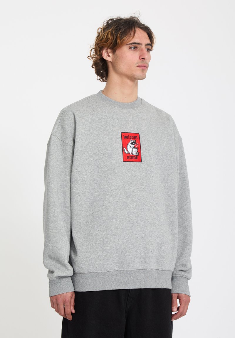 Joven con sudadera gris con un parche del logo rojo de Volcom Stone, de pie con expresión neutral contra un fondo liso.