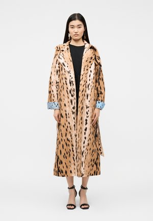 Donna in piedi che indossa un lungo cappotto beige a pois neri con dettagli azzurro chiaro sui polsini, vestito nero e sandali neri con tacco alto.