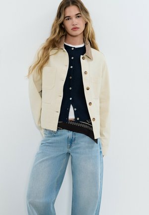 Mujer joven con cabello largo y rubio, vistiendo una chaqueta beige abotonada sobre un cárdigan azul marino y vaqueros de pierna ancha color azul claro.