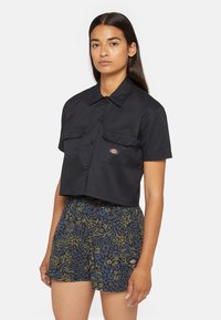 Schwarzes, gekürztes kurzärmliges Shirt mit zwei Vordertaschen, glatter Textur und Markennamen-Patch. Kombiniert mit gemusterten Shorts in Blau und Grün.