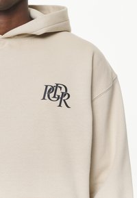 Sudadera beige hecha de una tela suave con un logo circular en gris oscuro. Tiene un corte relajado, con una capucha amplia y puños acanalados.