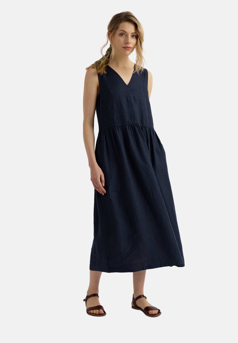 Jeune femme avec une tresse lâche portant une robe midi sans manches bleu marine à col en V et des sandales marron à brides à la cheville, debout sur un fond blanc.