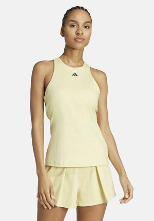 Femme portant un haut de sport sans manches jaune clair et un short plissé assorti, debout avec une expression neutre sur un fond blanc.