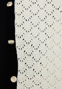 Cardigan tricotat cu un design din dantelă albă pe față, cu un contur solid negru și butoane aurii pentru închidere.