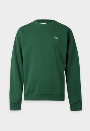 Homme portant un sweat-shirt vert et un pantalon de survêtement assorti avec des baskets blanches sur fond blanc.