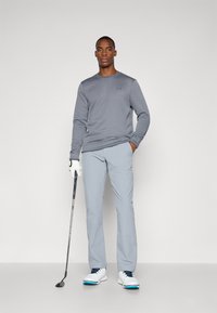 Gray long-sleeve shirt, light gray pants, white golf shoes with blue accents, holding a golf club, standing against a plain background.