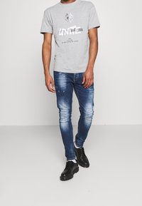 Camiseta de algodón gris con estampado gráfico, combinada con vaqueros azules desgastados y zapatos negros con cordones. Los vaqueros presentan secciones rasgadas y áreas desvaídas.