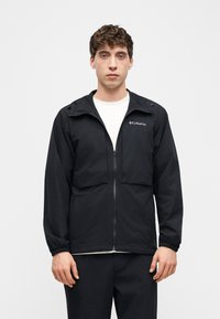 Νεαρός άνδρας που φοράει μαύρο εξωτερικό μπουφάν Columbia με φερμουάρ πάνω από λευκό πουκάμισο, στέκεται μπροστά σε απλό ανοιχτό φόντο, κοιτάζοντας προς τα εμπρός.