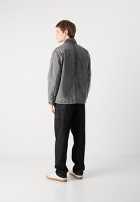 Grå denimjakke med krage, avslappet passform, synlige sømmer og knappede mansjetter, sammen med mørke bukser og hvite sneakers.
