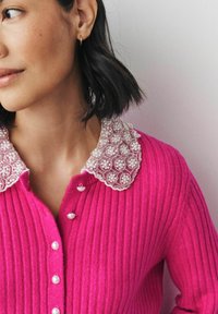 Helle pinke, gerippte Strickjacke mit einem gewellten Kragen, der mit weißen Perlen verziert ist und Perlmuttknöpfen. Kurze Ärmel runden das Design ab.