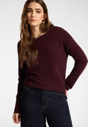 Ung kvinde med langt brunt hår iført en ribbet mørk lilla sweater, mørke jeans, guldkæde halskæde og guldhoop øreringe.