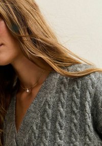 Cardigan grigio a maglia con scollo a V profondo, abbinato a una delicata collana d'oro con una singola perla. Texture morbida e vestibilità comoda.