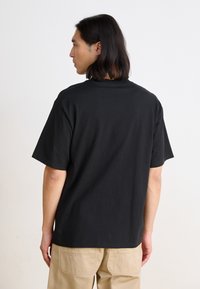 Black cotton t-shirt with short sleeves, crew neck, and a relaxed fit. No visible patterns or logos. Plain design, worn with beige pants.