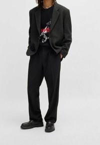 Blazer oversized nero in tessuto texture, indossato sopra una t-shirt grafica nera con un cavallo e testo rosso, abbinato a pantaloni neri larghi e scarpe nere.