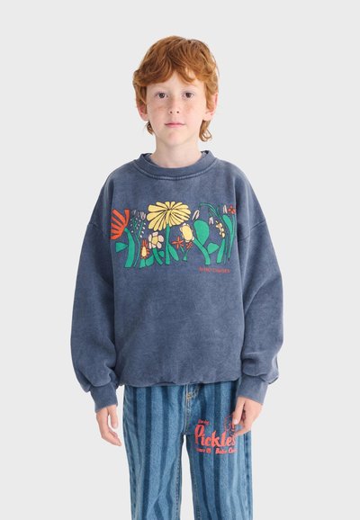 Un niño pelirrojo está de frente, lleva una sudadera azul con un diseño floral colorido y vaqueros azules a rayas con texto rojo.