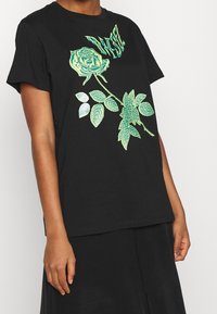 Camiseta de algodón negra con un gráfico floral verde de una rosa y hojas. Cuello redondo y mangas cortas. Corte simple y ajuste relajado.