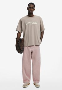 Khaki oversize tričko s grafikou "DESIGUAL"; růžové široké kalhoty; nese hnědou pruhovanou kabelku; béžové tenisky.