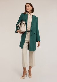 Cappotto verde in lana con una vestibilità su misura; maglione color crema con coulisse; pantaloni beige cropped; scarpe con tacco nude; borsa bianca.