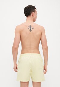 Shorts de bain pour hommes de couleur jaune clair, taille élastique, poche arrière, texture lisse, design sobre, portés par une personne avec un tatouage dans le dos.