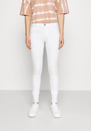 Person trägt enge weiße Jeans, weiße Plateau-Sneaker und ein kurzärmeliges, pfirsich-weißes Batik-Oberteil und steht vor einem schlichten hellen Hintergrund.