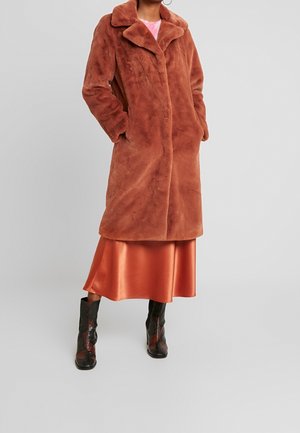 Kobieta ubrana w długi, rudy płaszcz z futra sztucznego, na pomarańczowej satynowej spódnicy i czarnych botkach na obcasie z wzorem skóry węża.