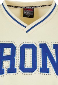 Cream-colored jersey with a V-neck, featuring blue embroidered lettering "IRON" embellished with silver studs along the text.