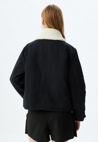 Schwarze Jacke mit einem weißen, flauschigen Kragen, lockerer Passform, weichem Material und sichtbaren Nähten auf der Rückseite. Getragen über schwarzen Shorts.