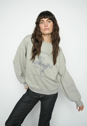 Frau mit langen welligen Haaren, die einen grauen Sweatshirt mit Blumenmuster und Schriftzug trägt, kombiniert mit dunklen Jeans, posiert vor einem einfarbigen weißen Hintergrund.