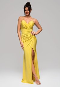 Robe de soirée jaune avec un décolleté en V profond, fronces sur le corsage et une fente haute. Réalisée en tissu lisse, accompagnée de sandales métalliques.