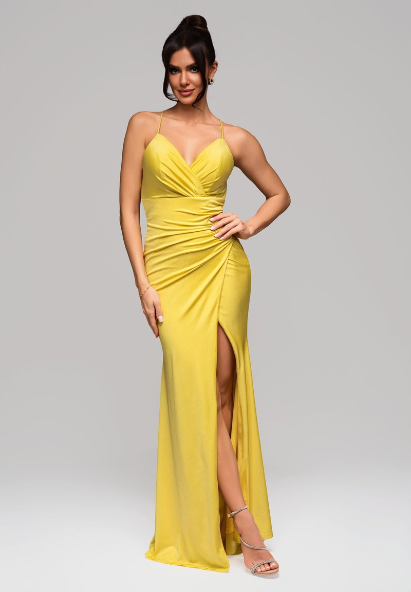 Robe de soirée jaune avec un décolleté en V profond, fronces sur le corsage et une fente haute. Réalisée en tissu lisse, accompagnée de sandales métalliques.