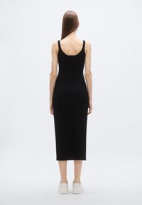 Čierne priliehavé midi šaty s širokými ramienkami a výstrihom na chrbte, vyrobené z elastického materiálu. Nosia sa s bielymi teniskami, hladká textúra.